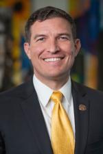 Portrait of a smiling man in a dark suit and yellow tie, wearing a lapel pin, against a colorful blurred background.