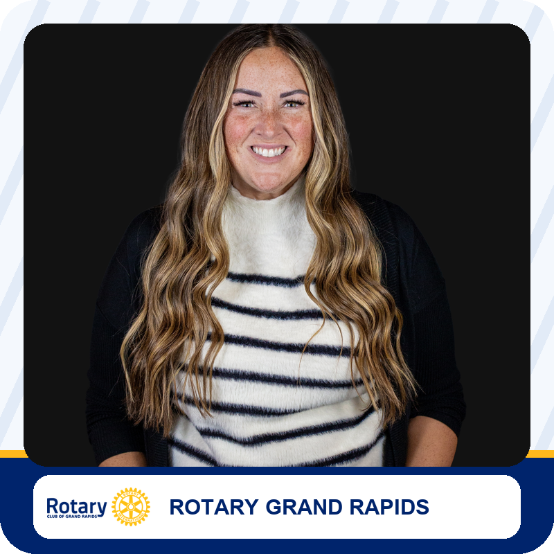 Portrait of a smiling woman with long blonde hair in a white and navy striped sweater against a dark background for Rotary Grand Rapids.
