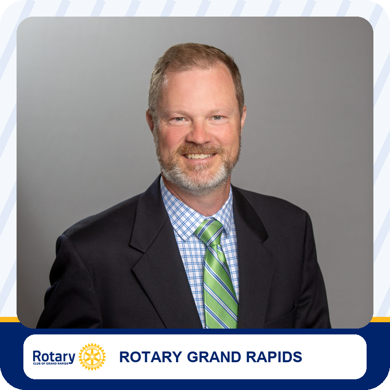 Portrait of a smiling man in a dark suit, blue check shirt, and green striped tie against a gray backdrop, used for Rotary Grand Rapids branding.