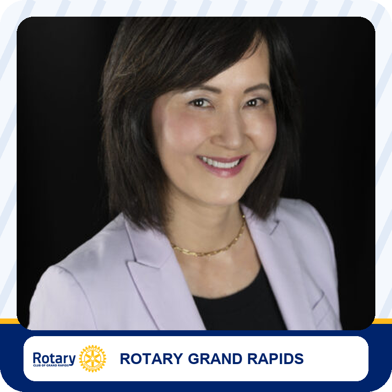 Headshot of a smiling woman in a lavender blazer with a gold necklace, posed against a dark background for Rotary Grand Rapids.