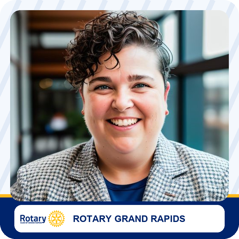 Smiling person in a checkered blazer inside a modern space, Rotary Grand Rapids banner below.
