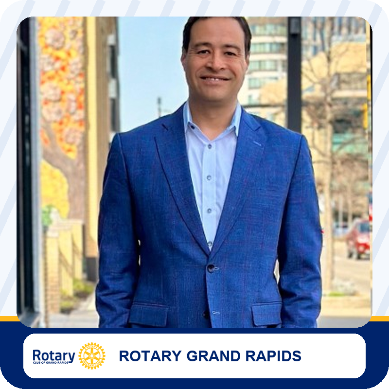Smiling man in a blue suit standing on a city sidewalk, Rotary Grand Rapids branding below.