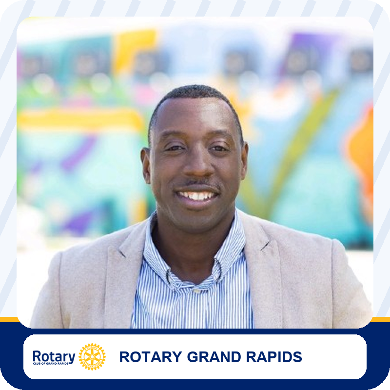 Smiling man in a beige blazer and striped shirt posing outdoors, Rotary Grand Rapids branding at bottom.