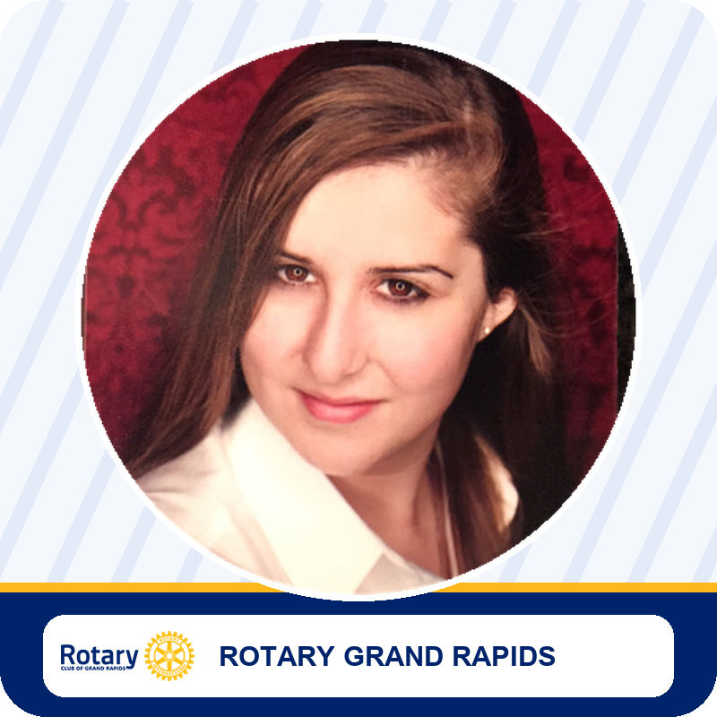 Head-and-shoulders portrait of a woman with brown hair, white shirt, in a circular frame on a Rotary Grand Rapids banner background.